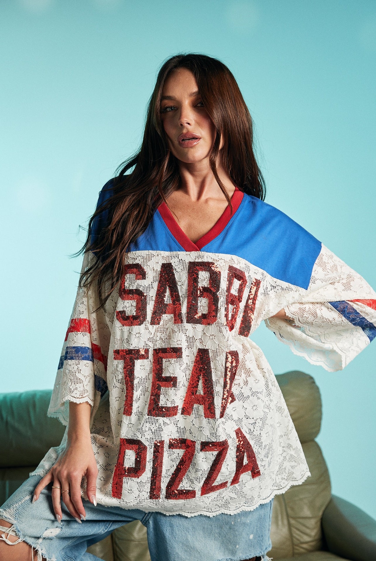 Woman wearing a loose lace top with bold red-sequined text ‘Sabbi Team Pizza,’ paired with distressed denim pants, posing against a light blue background.