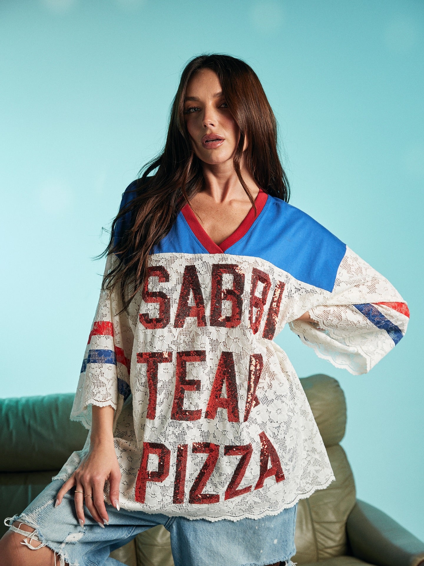 Woman wearing a loose lace top with bold red-sequined text ‘Sabbi Team Pizza,’ paired with distressed denim pants, posing against a light blue background.