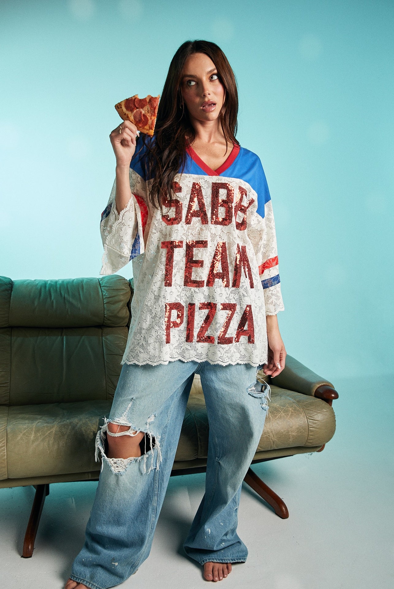 Woman holding a slice of pepperoni pizza while wearing oversized lace Sabbi Team Pizza top with red lettering and distressed wide-leg jeans.