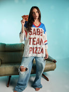 Woman holding a slice of pepperoni pizza while wearing oversized lace Sabbi Team Pizza top with red lettering and distressed wide-leg jeans.