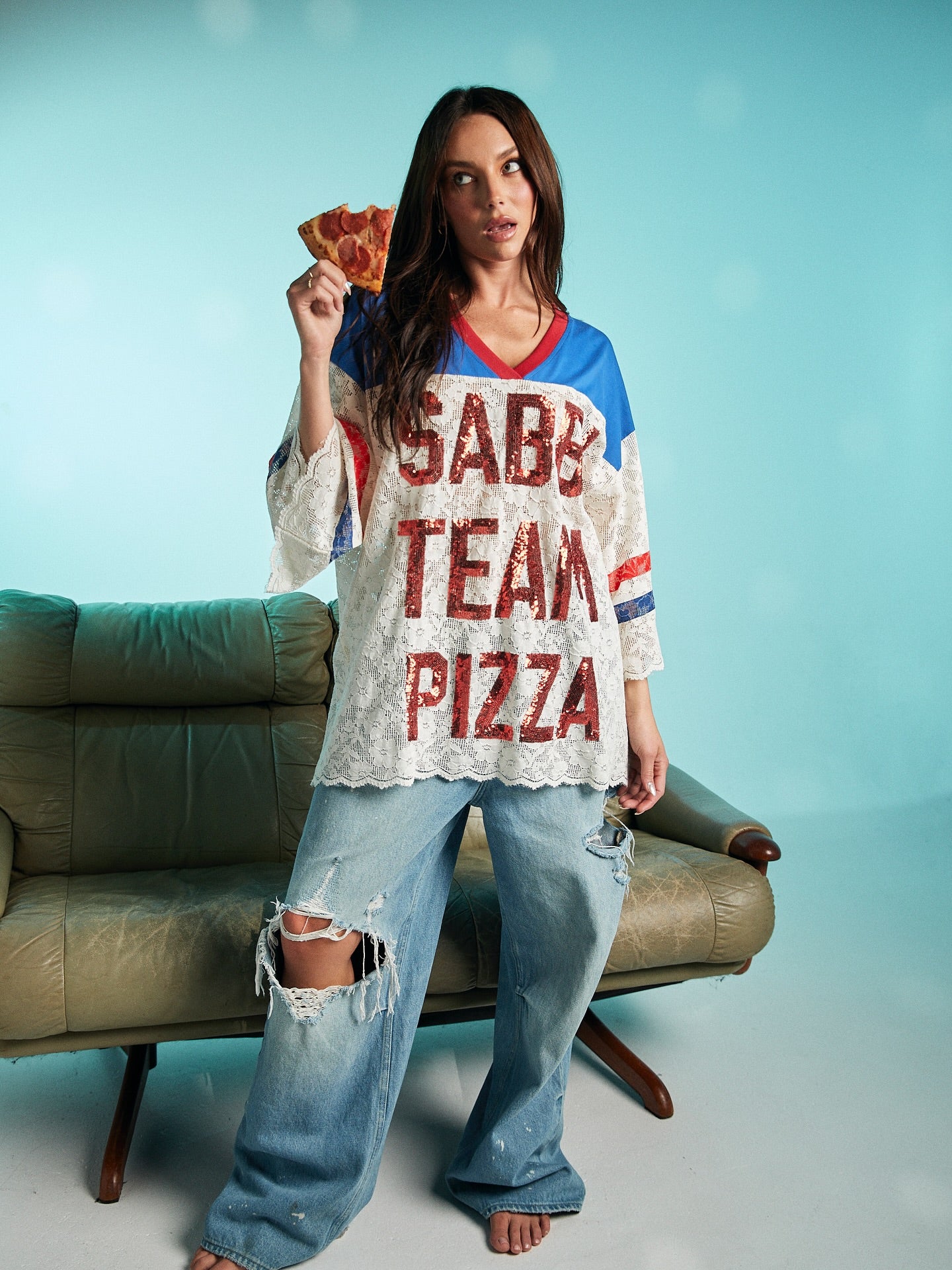Woman holding a slice of pepperoni pizza while wearing oversized lace Sabbi Team Pizza top with red lettering and distressed wide-leg jeans.