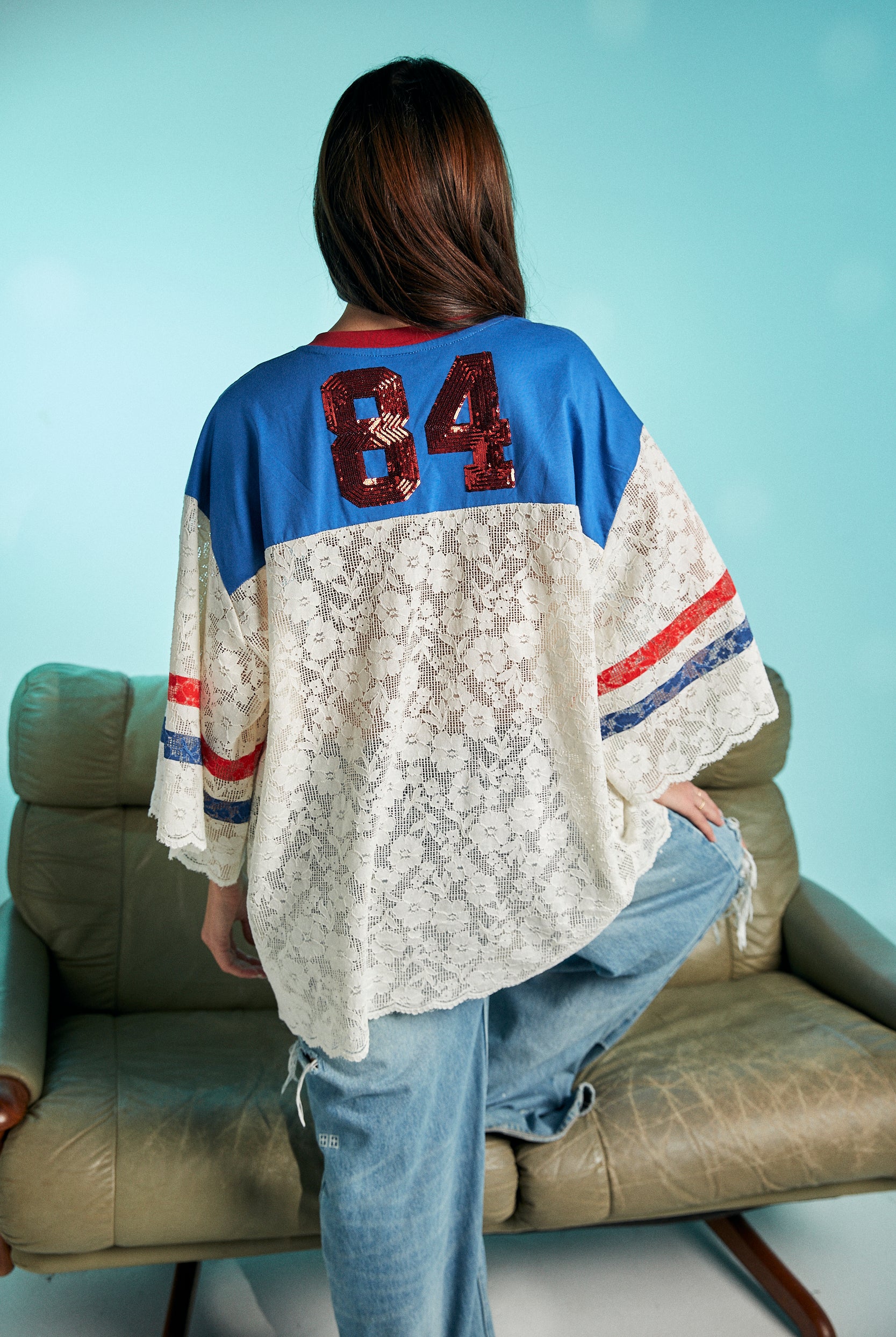 Back view of woman wearing oversized lace top with blue shoulders, red sequined number 84, and red and blue striped sleeves, styled with ripped denim jeans.